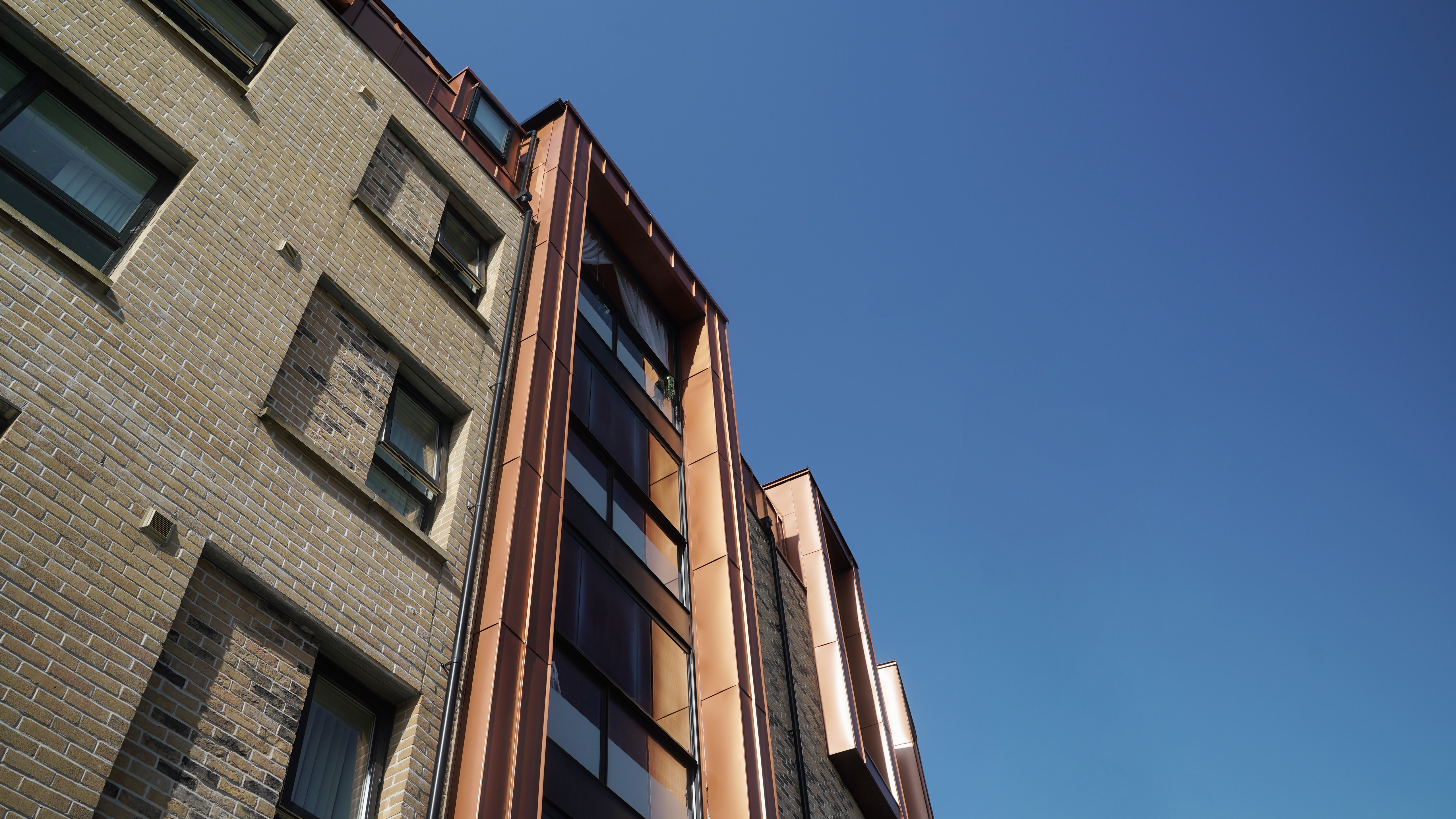 Perspective view of the façade of a modern residential development on Nethan Street in Glasgow, clad with FALZONAL in New Copper. The metallic façade elements catch and reflect the light, emphasising the shape of the building and providing an impressive visual contrast to the traditional brick architecture. The PREFA aluminium product not only provides an appealing aesthetic, but also promises durability and low maintenance.