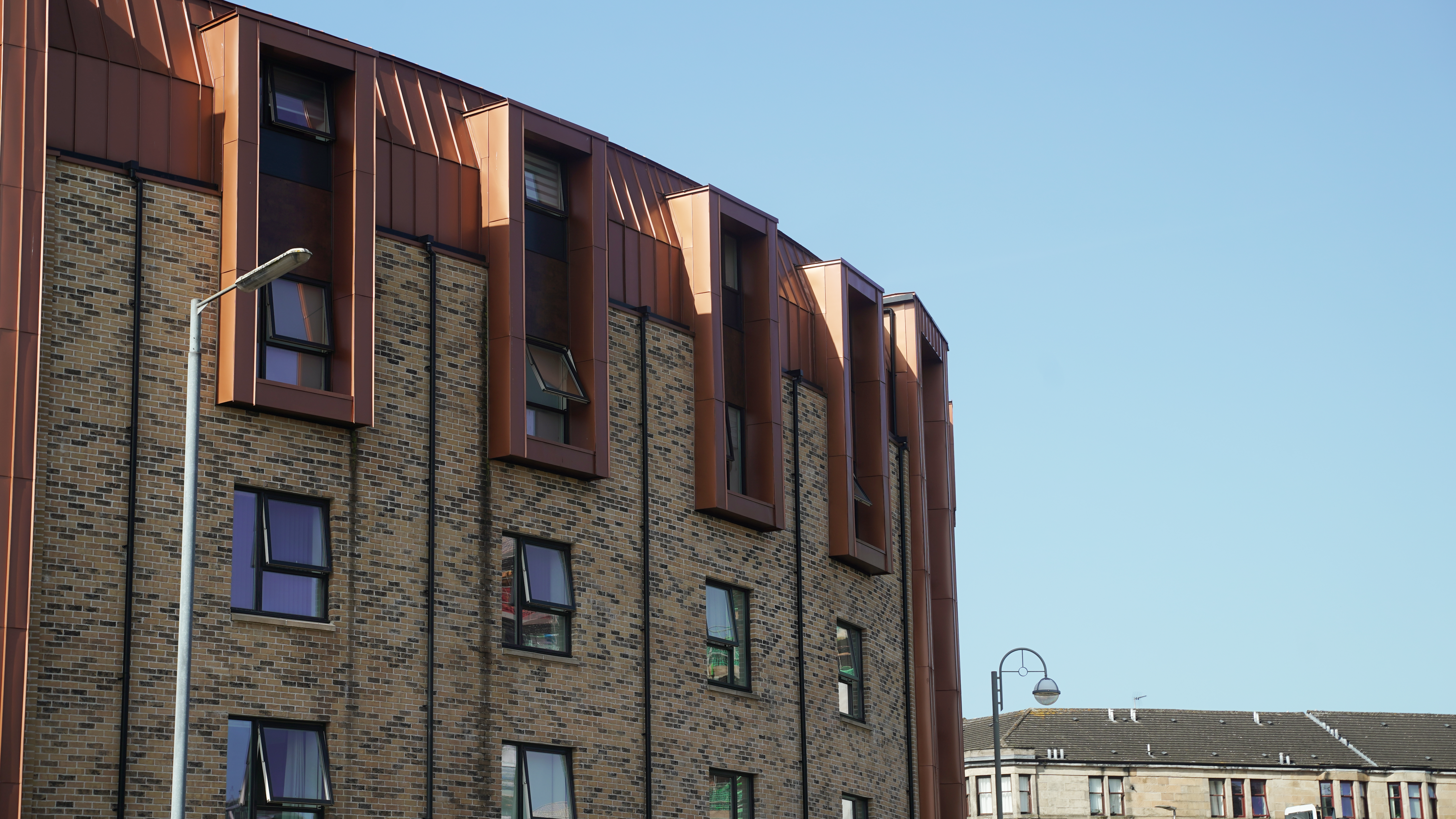 The façade of a residential development on Nethan Street in Glasgow, accentuated by the tasteful use of FALZONAL in New Copper. The PREFA aluminium product gives the building envelope a vibrant and inviting appearance through its reflection of sunlight. The combination of the traditional brick structure with the modern, copper-coloured accents of the FALZONAL conveys an elegant symbiosis of old and new and highlights the architecture in this urban context.
