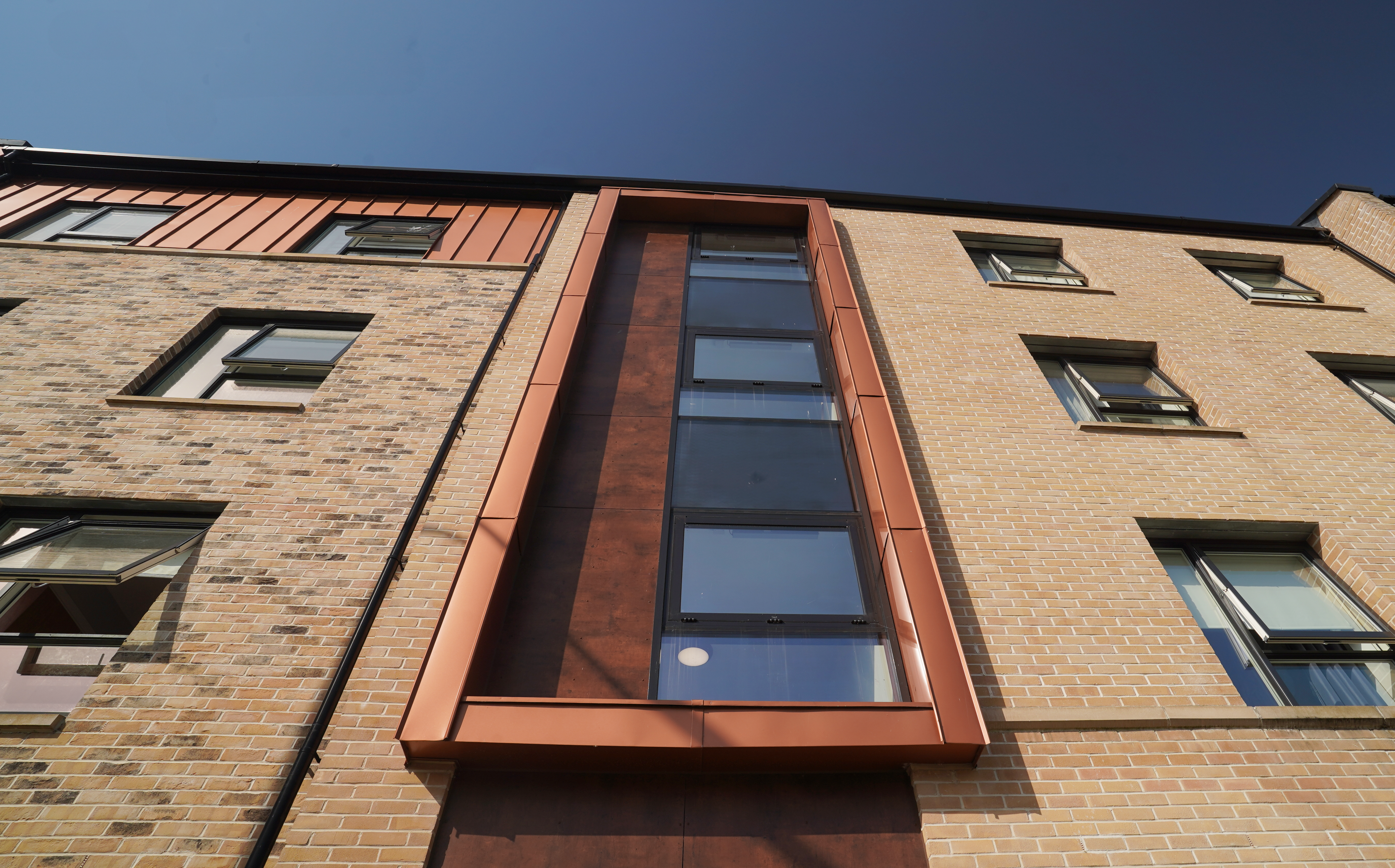 Detailed view of the façade of a residential building on Nethan Street in Glasgow, characterised by the cladding with FALZONAL in new copper. The copper-coloured aluminium cladding around the window sections sets modern accents against the classic brick façade and shows the interplay of innovative materials and traditional design. The high-quality aluminium product from PREFA underlines the aesthetics of the building and at the same time ensures its durability and weather resistance.