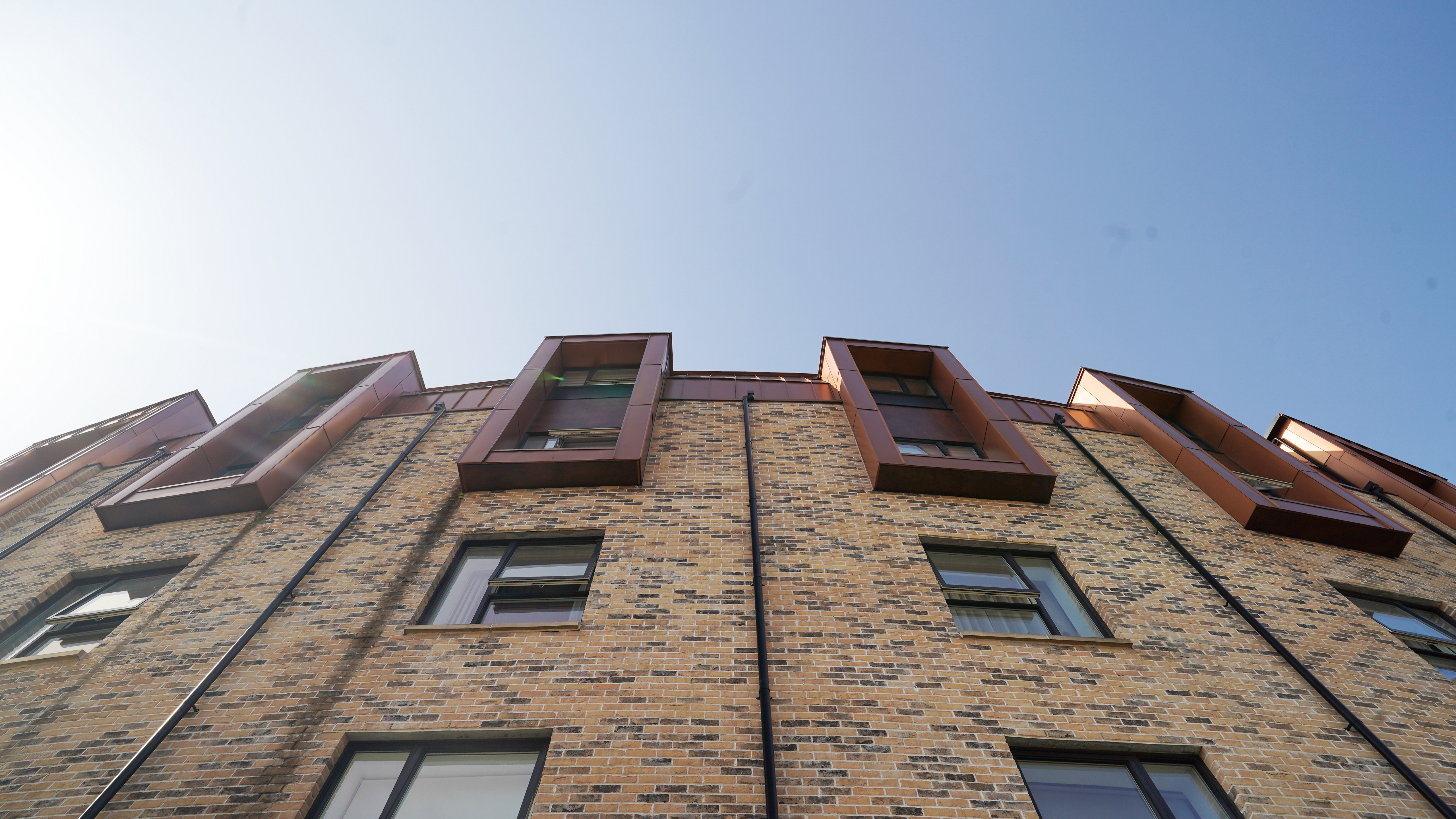 An upward view of the façade of a residential development on Nethan Street in Glasgow, which features the use of FALZONAL in New Copper, a durable and robust PREFA aluminium product. The vertical alignment of the windows, framed with the copper-coloured aluminium, breaks up the horizontal articulation of the brick wall and gives the building a dynamic and modern look. The quality and colouring of the FALZONAL blends seamlessly into the urban architecture while offering a durable and aesthetic solution for modern residential complexes. Black downpipes for roof drainage have been placed between the windows, emphasising the curvature of the building from this perspective.